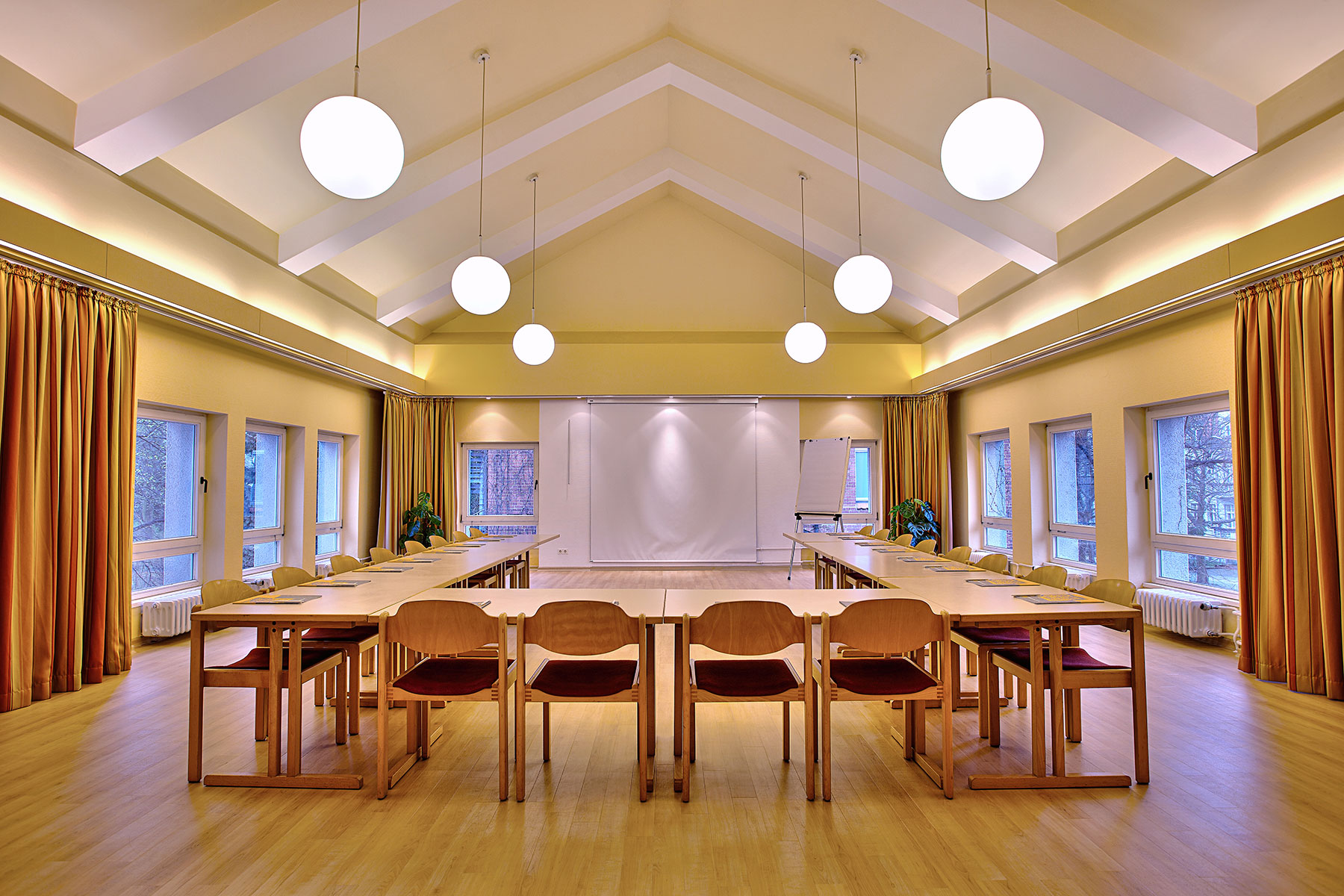 Foto von einem Konferenzraum, die Tische sind eingedeckt für 20 Personen mit Blöcke, rechts und links gibt es Fenster mit Vorhängen, an der Front hängt eine Leinwand