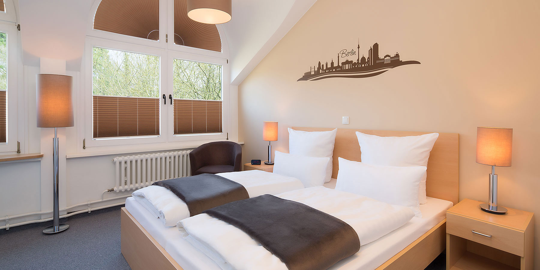 Doppelzimmer im Neubau mit zwei Betten und Beistelltisch. Ein Sessel steht neben dem Bett, ein Stuhl am Schreibtisch, der TV hängt an der Wand und es gibt einen Kleiderschrank. Das Fenster ist links neben dem Bett. Der Boden ist mit einem Teppich ausgelegt. Es ist möglich die Fenster zu verdunkeln.