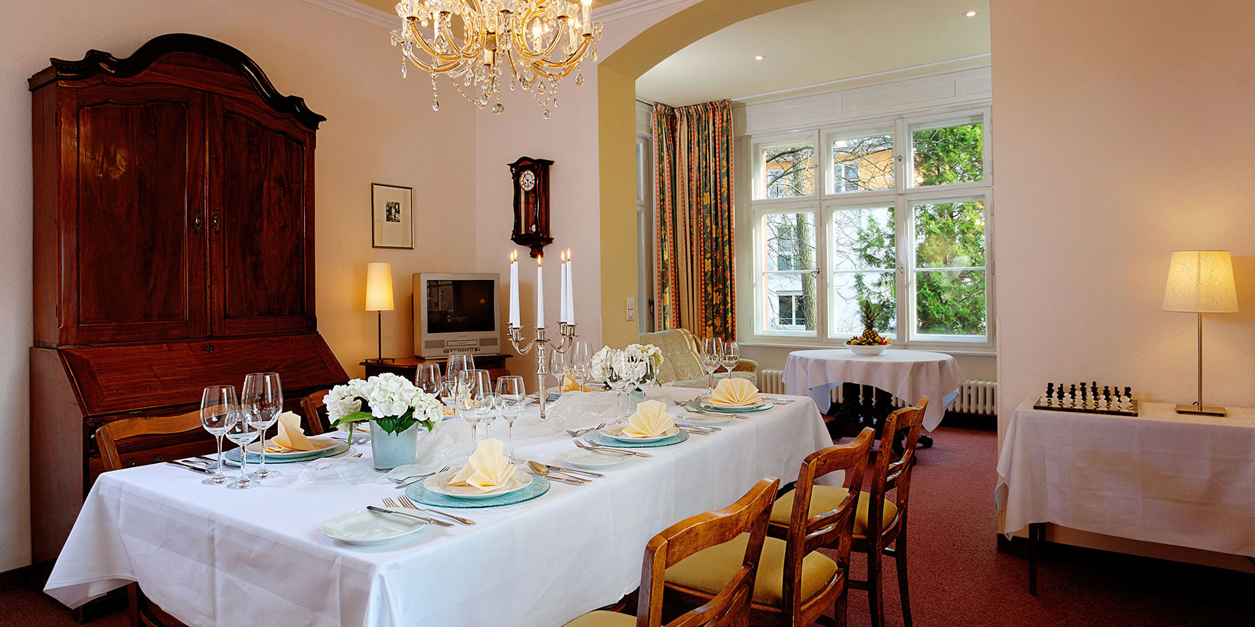 Tagungsraum Jubilate, ein Tisch ist feierlich eingedeckt für 6 Gäste, Weingläser, Teller, Serviette, Besteck für 4 Gänge, Blumengesteck, Tischdecke, Kerzenständer rechts neben dem Tisch ist ein Fenster zum Garten raus, links hinter dem Tisch steht ein Sekretär und TV