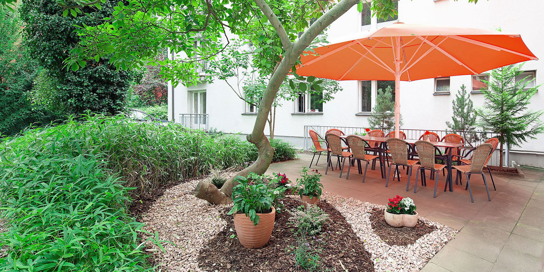 Ansicht von der Terrasse am Frühstücksraum, es ist sehr grün und die Sonne scheint, es stehen zwei Tische mit Stühlen darauf, links davor ist ein Beet bepflanzt