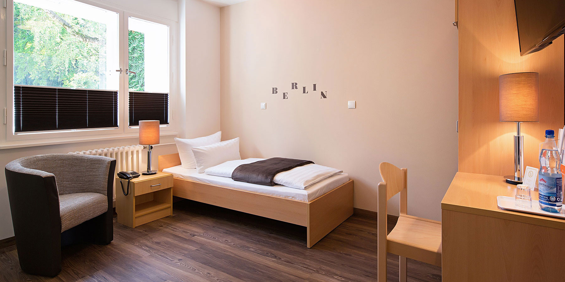 Einzelzimmer im Neubau mit einem Bett und Beistelltisch. Dieses Zimmer ist Barrierefrei. Ein Sessel steht neben dem Bett, ein Stuhl am Schreibtisch, der TV hängt an der Wand und es gibt einen Kleiderschrank. Das Fenster ist hinter dem Bett. Der Boden ist Vinyl ausgelegt. Es ist möglich die Fenster zu verdunkeln.