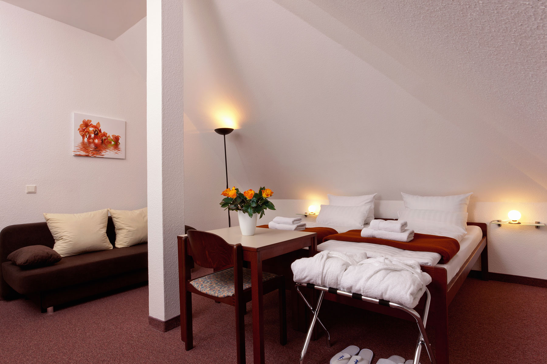 Doppelzimmer mit zwei Betten und Beistelltisch. Eine Couch steht links daneben, diese kann man Aufbetten, der TV hängt an der Wand und es gibt einen Kleiderschrank. Das Fenster ist links neben der Couch. Der Boden ist mit einem Teppich ausgelegt. Es ist möglich die Fenster zu verdunkeln.