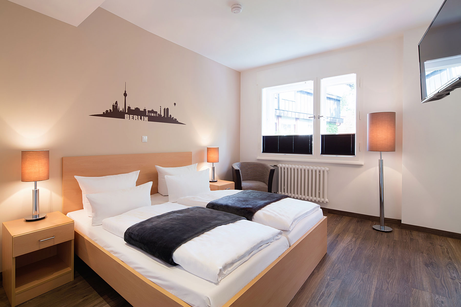 Doppelzimmer im Neubau mit zwei Betten und Beistelltisch. Dieses Zimmer ist Barrierefrei. Ein Sessel steht neben dem Bett, ein Stuhl am Schreibtisch, der TV hängt an der Wand und es gibt einen Kleiderschrank. Das Fenster ist rechts neben dem Bett. Der Boden ist Vinyl ausgelegt. Es ist möglich die Fenster zu verdunkeln.