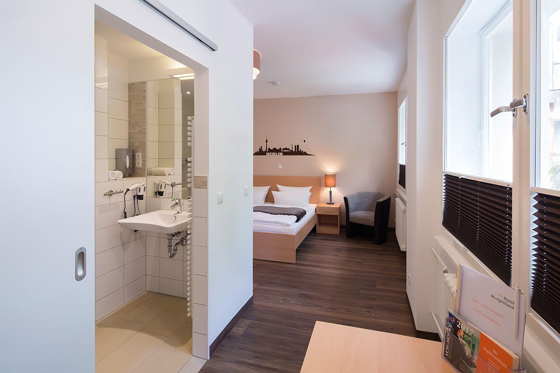 Doppelzimmer im Neubau mit zwei Betten und Beistelltisch. Dieses Zimmer ist Barrierefrei. Ein Sessel steht neben dem Bett, ein Stuhl am Schreibtisch, der TV hängt an der Wand und es gibt einen Kleiderschrank. Das Fenster ist rechts neben dem Bett. Der Boden ist Vinyl ausgelegt. Es ist möglich die Fenster zu verdunkeln.