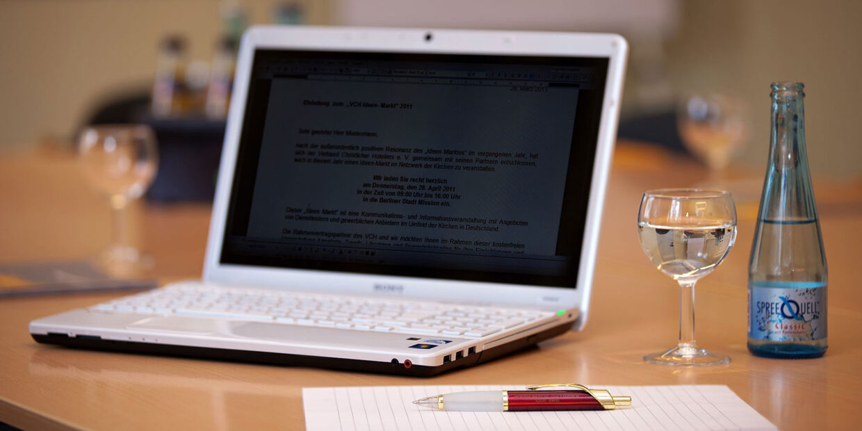 Detailfoto vom Seminarraum auf dem Tisch steht ein Notebook, rechts daneben eine Wasser und ein Glas, davor ein Block mit Stift