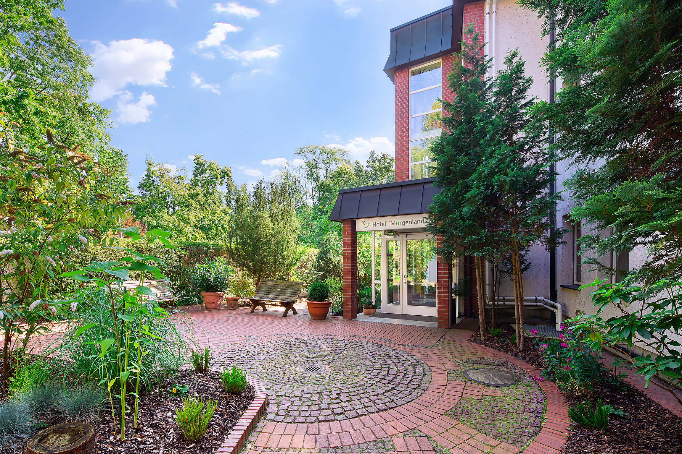 Außenansicht vom Hotel Morgenland, Haupteingang es ist sehr grün, die Sonne scheint, ein paar Wolken sind zusehen, links neben dem Haupteingang stehen zwei Bänke sowie Blumentöpfe, rechts daneben ist ein Beet bepflanzt