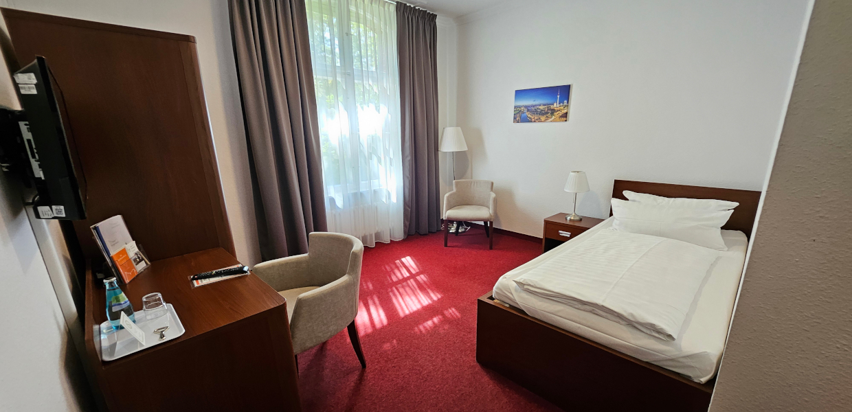 Einzelzimmer mit einem Bett und Beistelltisch. Ein Sessel steht am Schreibtisch, der TV hängt an der Wand und es gibt einen Kleiderschrank. Das Fenster ist rechts neben dem Schrank. Der Boden ist mit einem Teppich ausgelegt. Es ist möglich die Fenster zu verdunkeln.