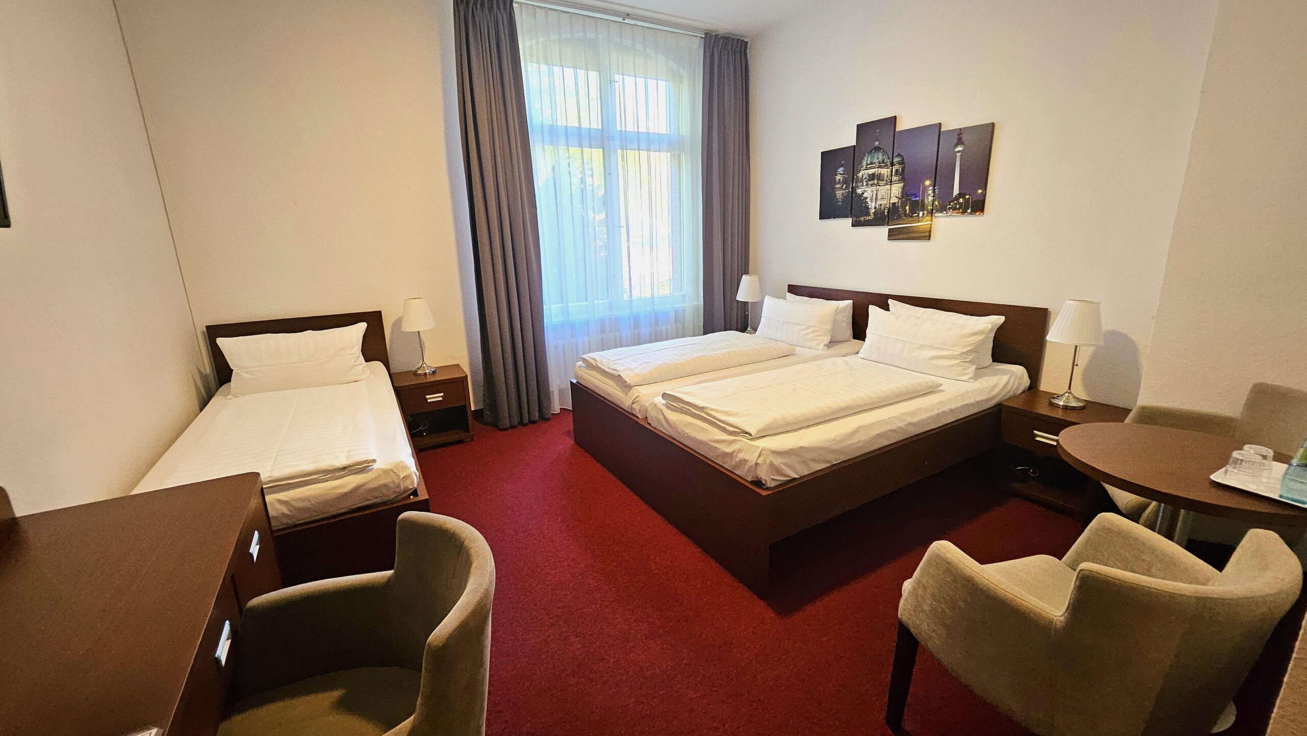 Dreibettzimmer mit drei Betten und Beistelltisch. Zwei Sessel steht am Tisch rechts, der TV hängt an der Wand und es gibt einen Kleiderschrank. Ein Stuhl steht am Schreibtisch links vor dem Einzelbett. Das Fenster ist links neben dem Doppelbett. Der Boden ist mit einem Teppich ausgelegt. Es ist möglich die Fenster zu verdunkeln.