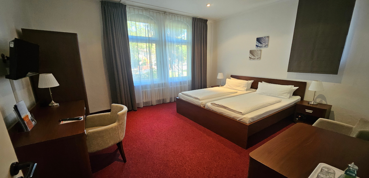 Doppelzimmer mit zwei Betten und Beistelltisch. Ein Sessel steht am Schreibtisch und einer rechts neben dem Bett, der TV hängt an der Wand und es gibt einen Kleiderschrank. Das Fenster ist links neben dem Bett. Der Boden ist mit einem Teppich ausgelegt. Es ist möglich die Fenster zu verdunkeln