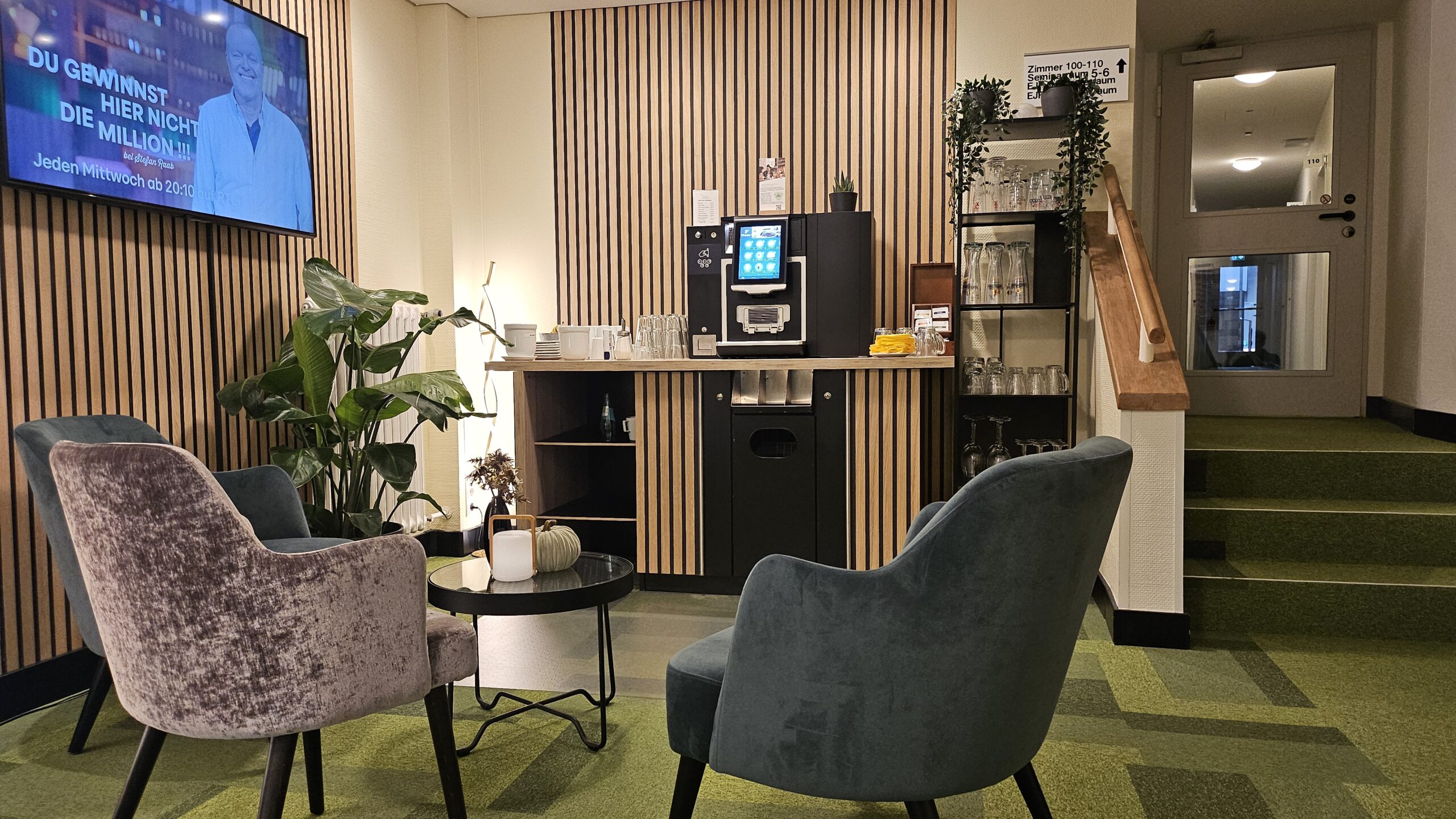 Coffeebar der neuen Lobby im Hotel Morgenland, es gibt drei bequeme Sessel, ein Tisch in der Mitte mit Blumen und Lichtern, im Hintergrund ist die Coffeebar zusehen und eine Regal mit Tassen und Gläsern, links an der Wand hängt ein TV