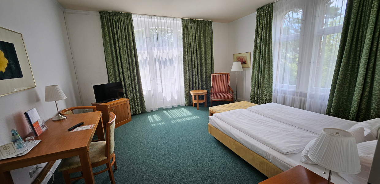 Doppelzimmer mit Balkon, einem Betten und Beistelltisch. Ein Stuhl steht am Schreibtisch links an der Wand, der TV steht auf einer Kommode neben der Balkontür. Der Balkon ist gegenüber vom Bett, rechts davon steht ein Sessel. Der Boden ist mit einem Teppich ausgelegt. Es ist möglich die Fenster zu verdunkeln.