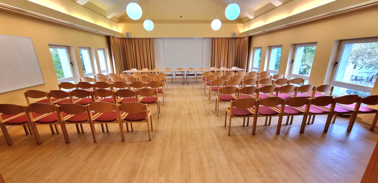 Konferenzraum mit Stuhlreihen für Sechzig Personen, vorne steht ein Podiumstisch für Zehn Personen, in der Mitte ist ein Gang frei bis zur Leinwand hinter dem Podiumstisch